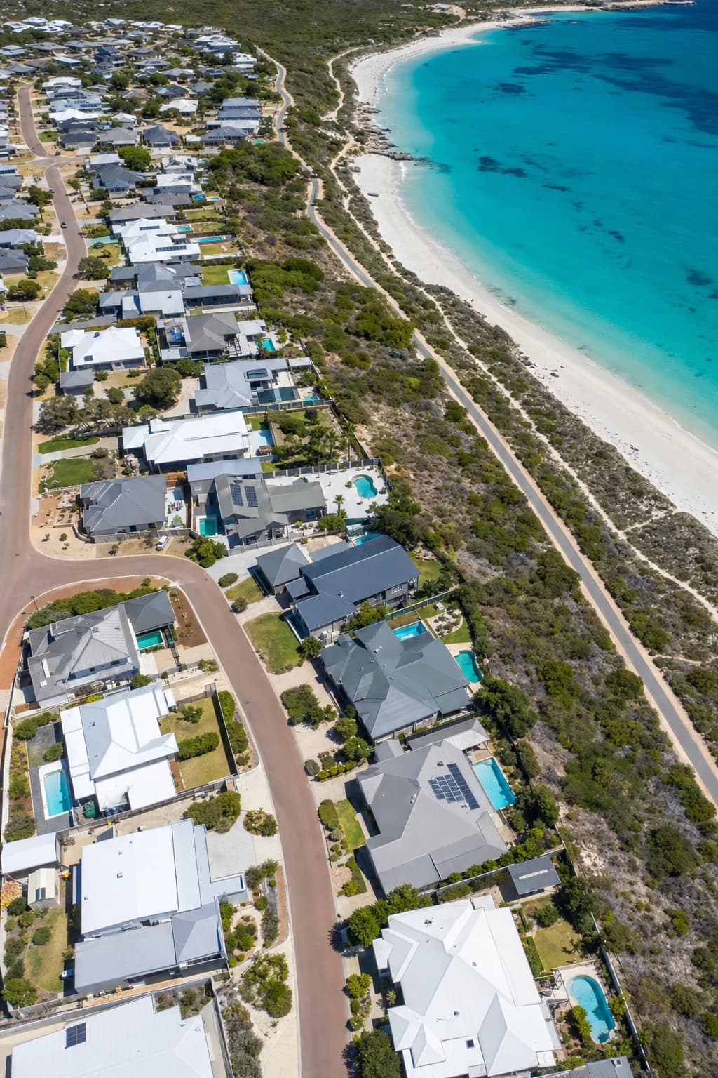 Aerial view of coastal homes with steel patios and pools beside a white sand beach and the bay