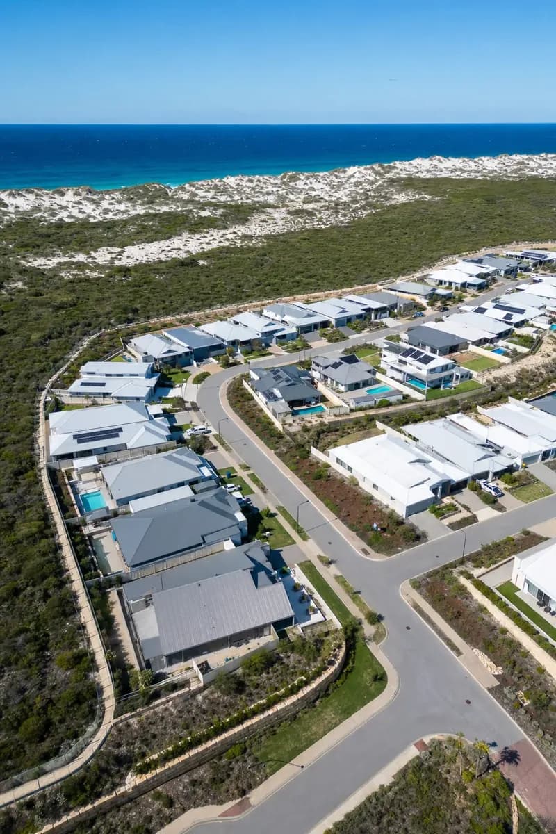 Aerial view of a new coastal estate in Yanchep with steel-roofed homes, pools, bushland and dunes by the ocean